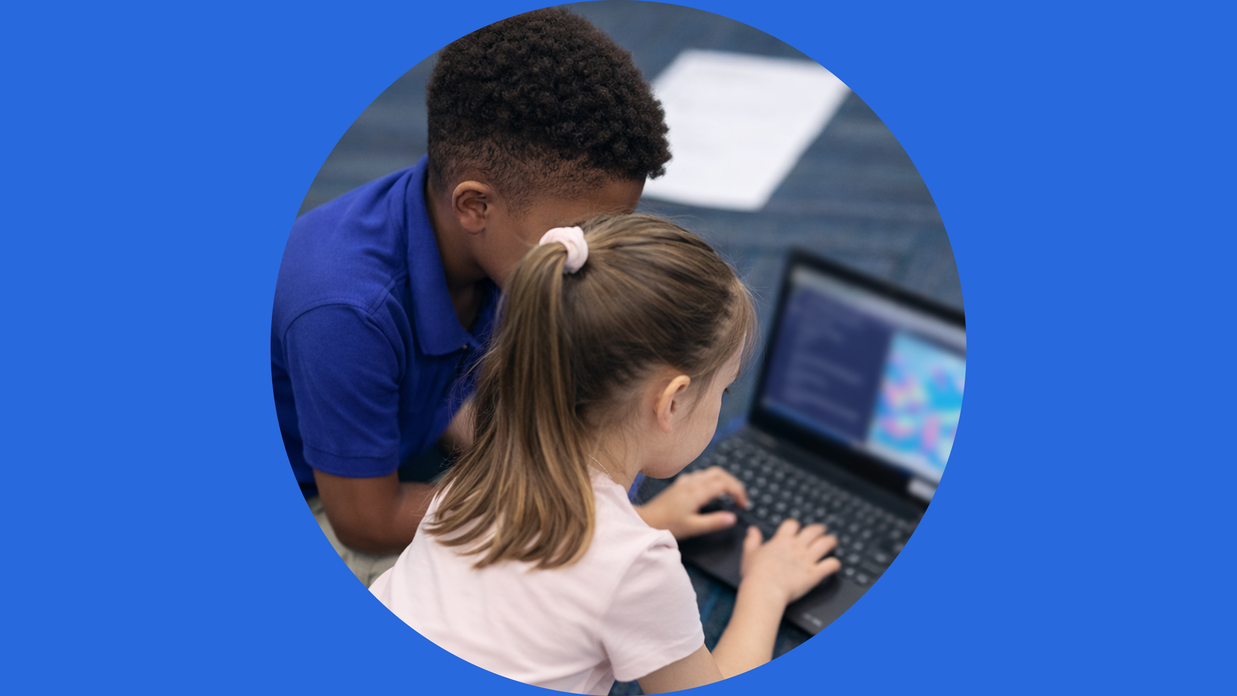 A young girl in a white shirt uses a computer keyboard while an instructor in a blue polo shirt observes and provides…