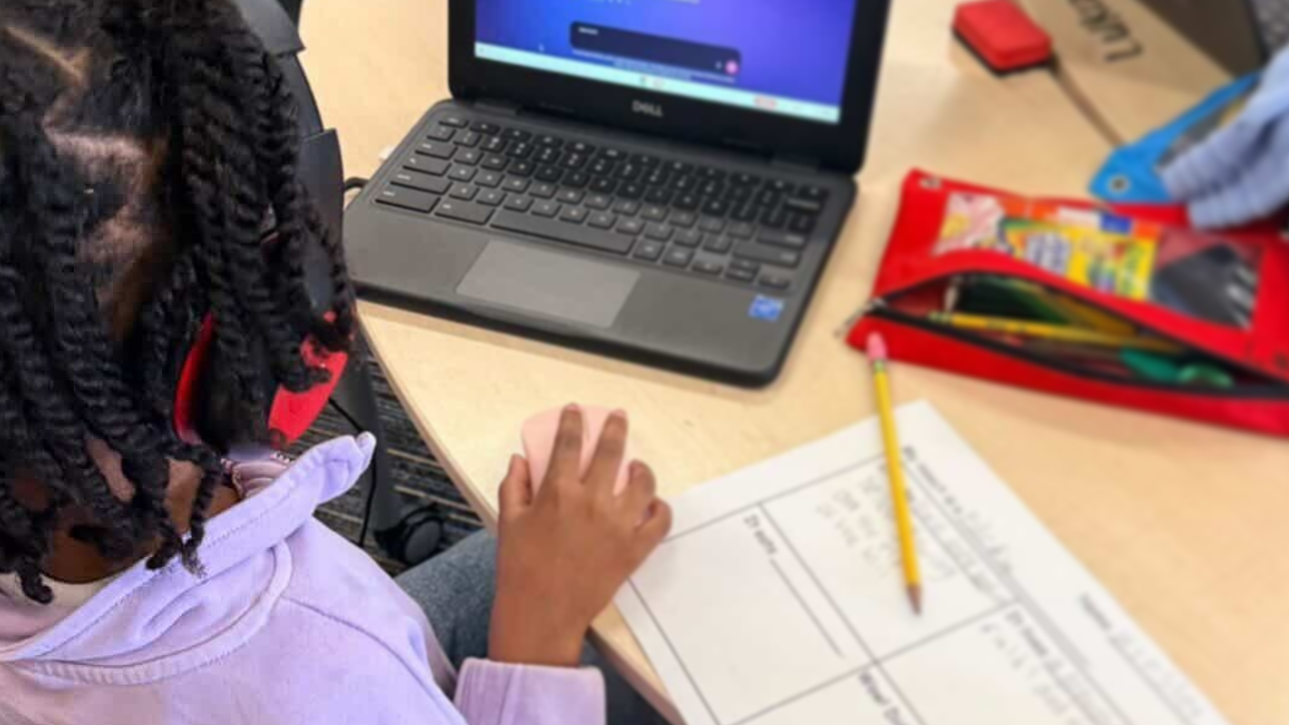 A student works at a wooden desk with a laptop, notebook, and colored pencils, engaged in schoolwork or homework.