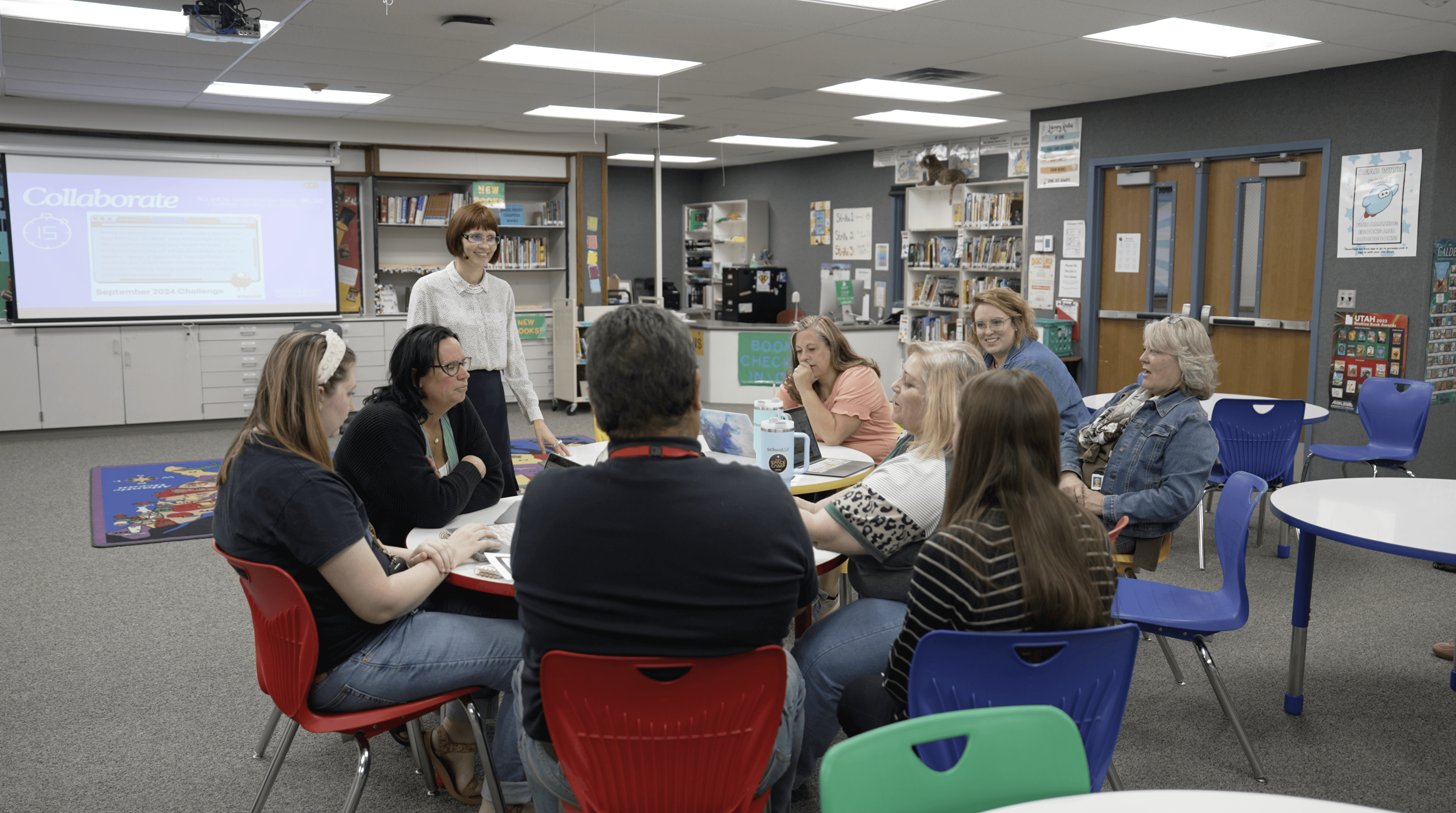 A group of educators sits in a circle during a collaborative discussion in a school library, with bookshelves and a…