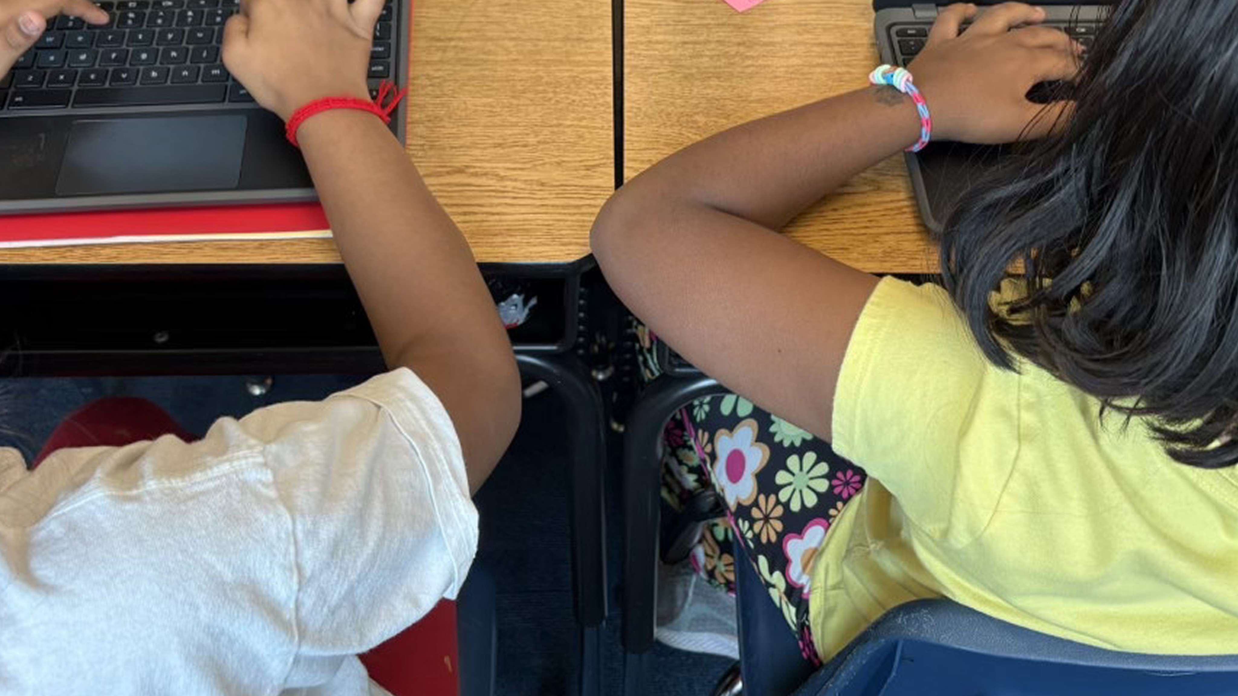 Two students sit at a wooden desk with laptops, wearing colorful bracelets. One wears a white shirt and the other a yellow…