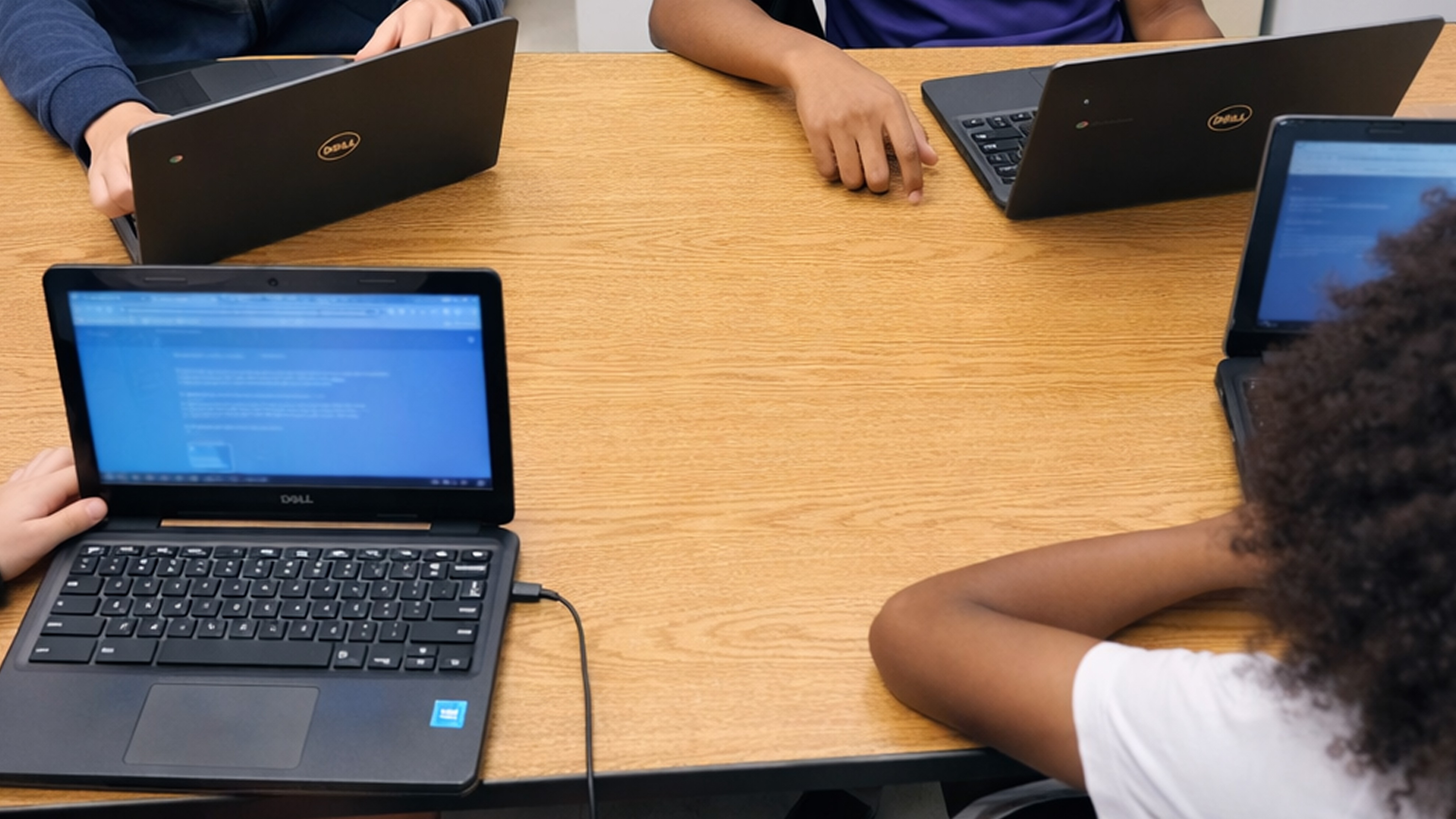 Multiple students work at a wooden table with laptops and netbooks displaying blue screens during a computer class or…