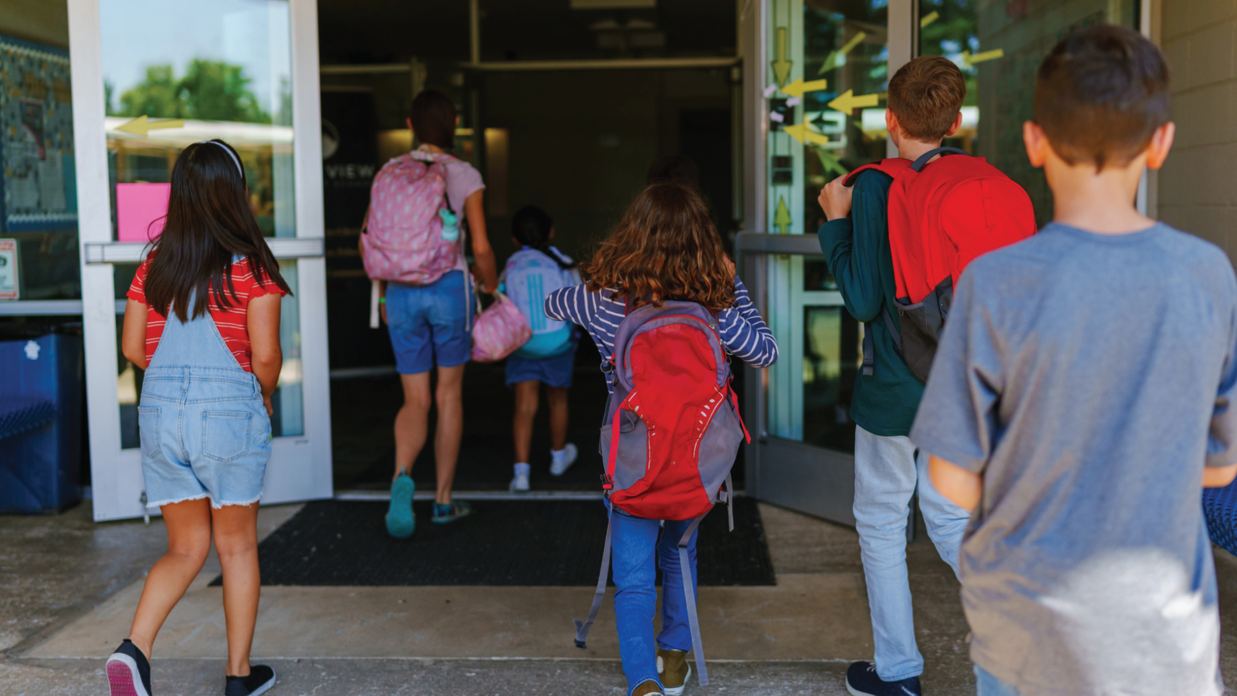 A group of elementary school children wearing backpacks entering a school building through glass doors.